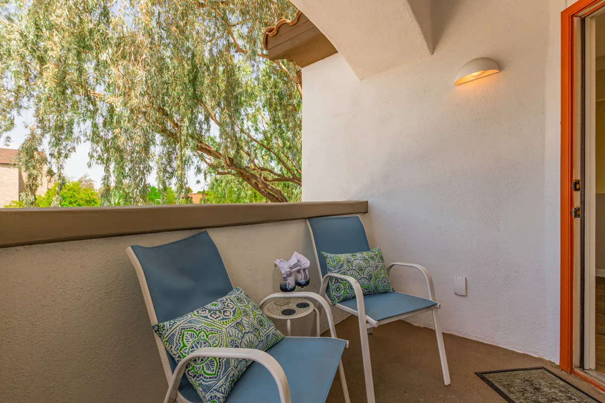 A serene private patio with two blue chairs, decorative pillows, a small table, and lush greenery outside. The space is shaded, offering a tranquil setting for enjoying morning coffee or quiet reading under the Arizona sky.