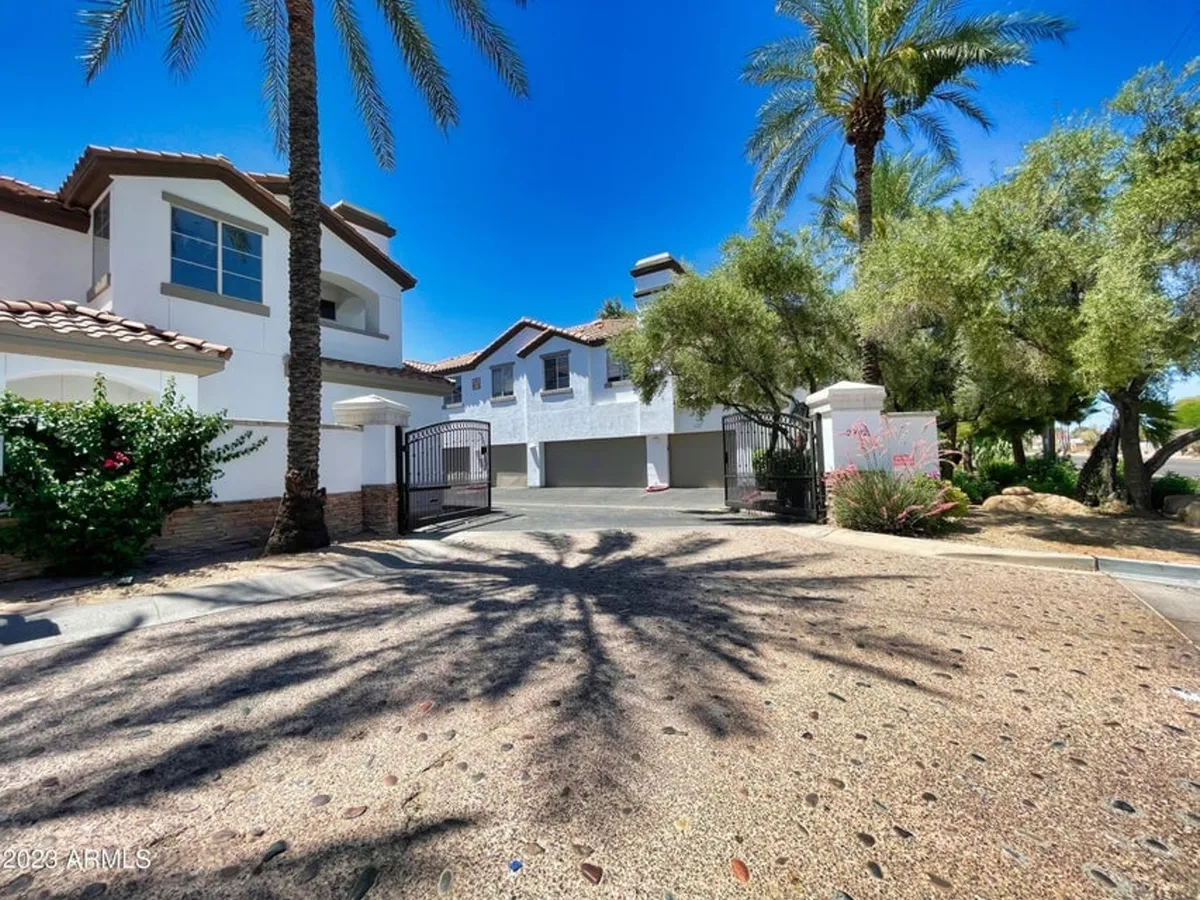 An inviting exterior showing a gated entrance framed by palm trees and a landscaped garden, with the house facade visible beyond. The gate represents security and exclusivity for residents and visitors alike.