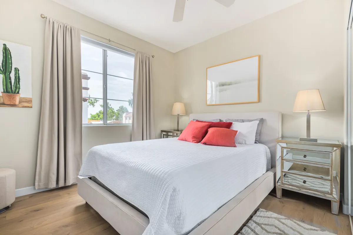 A cozy bedroom featuring a queen-sized bed with decorative pillows, a nightstand with a lamp, a window streaming with sunlight, and cactus-themed artwork. The room feels tranquil and is arranged for both comfort and aesthetic appeal.