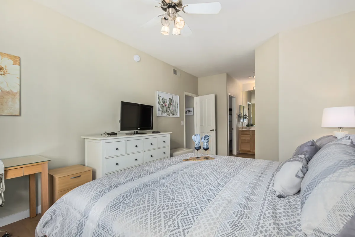 A serene bedroom space with a neatly made bed, a television for movie nights, modern artwork, and a desk by the window capturing gentle morning light.