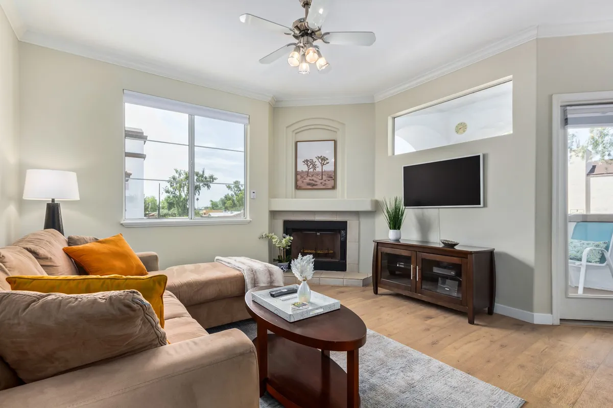 A bright, open living room featuring a plush sofa, working fireplace, TV, central coffee table, and large windows that fill the space with natural light. The vaulted ceilings amplify the sense of openness and airiness.