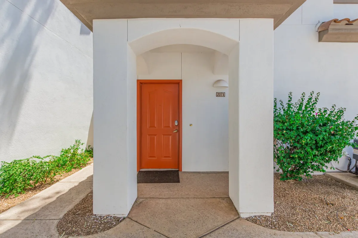The image captures the exterior of the townhome, focusing on a welcoming orange door framed by an arched entrance and lush green shrubbery. The inviting entryway hints at a peaceful, residential neighborhood and the sense of arrival that comes after exploring the lively local culture.