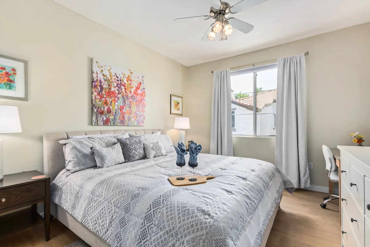 A serene bedroom with a neatly made bed adorned with decorative pillows, bright artwork above the headboard, and a window allowing natural light to fill the room. There's a small desk set up beside the bed, ideal for remote work or reading.