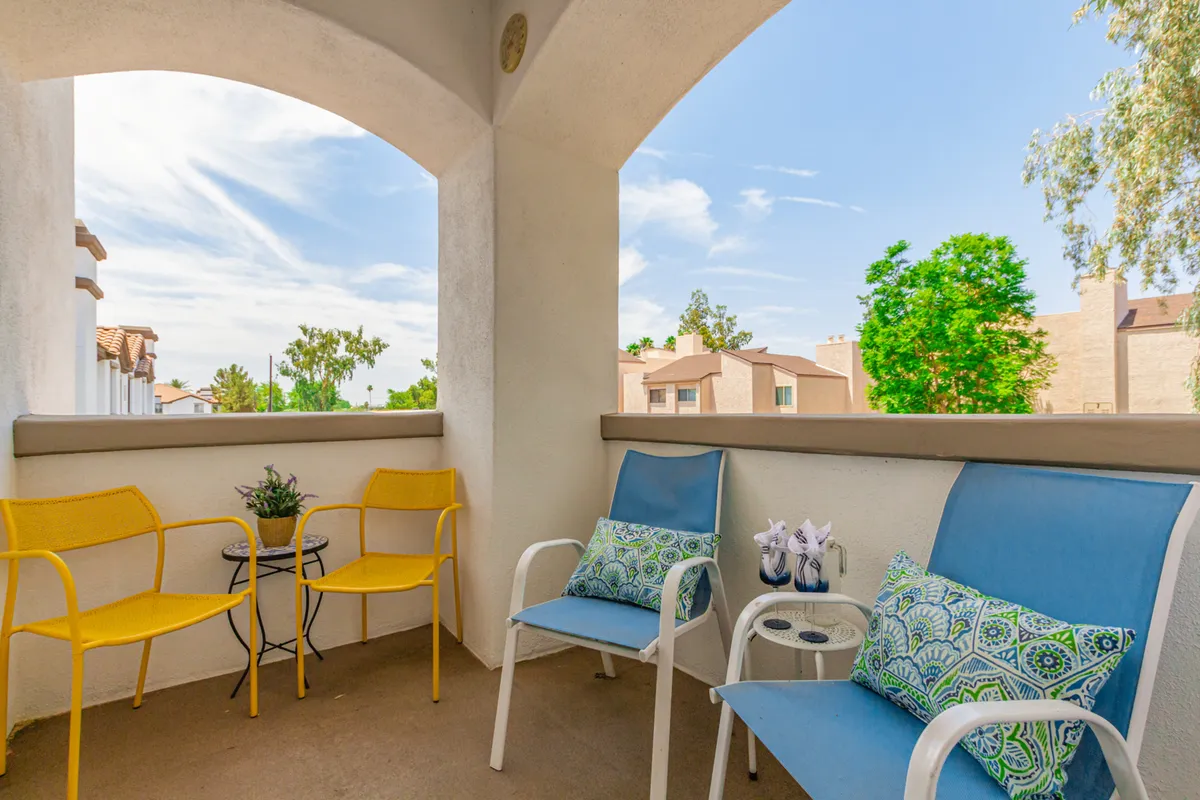 A cheerful outdoor patio scene featuring yellow and blue chairs with green cushions arranged around a small table, overlooking lush greenery and capturing the sense of relaxing and socializing outside. The area is bathed in natural light, suggesting either a tranquil morning or a pleasant evening.