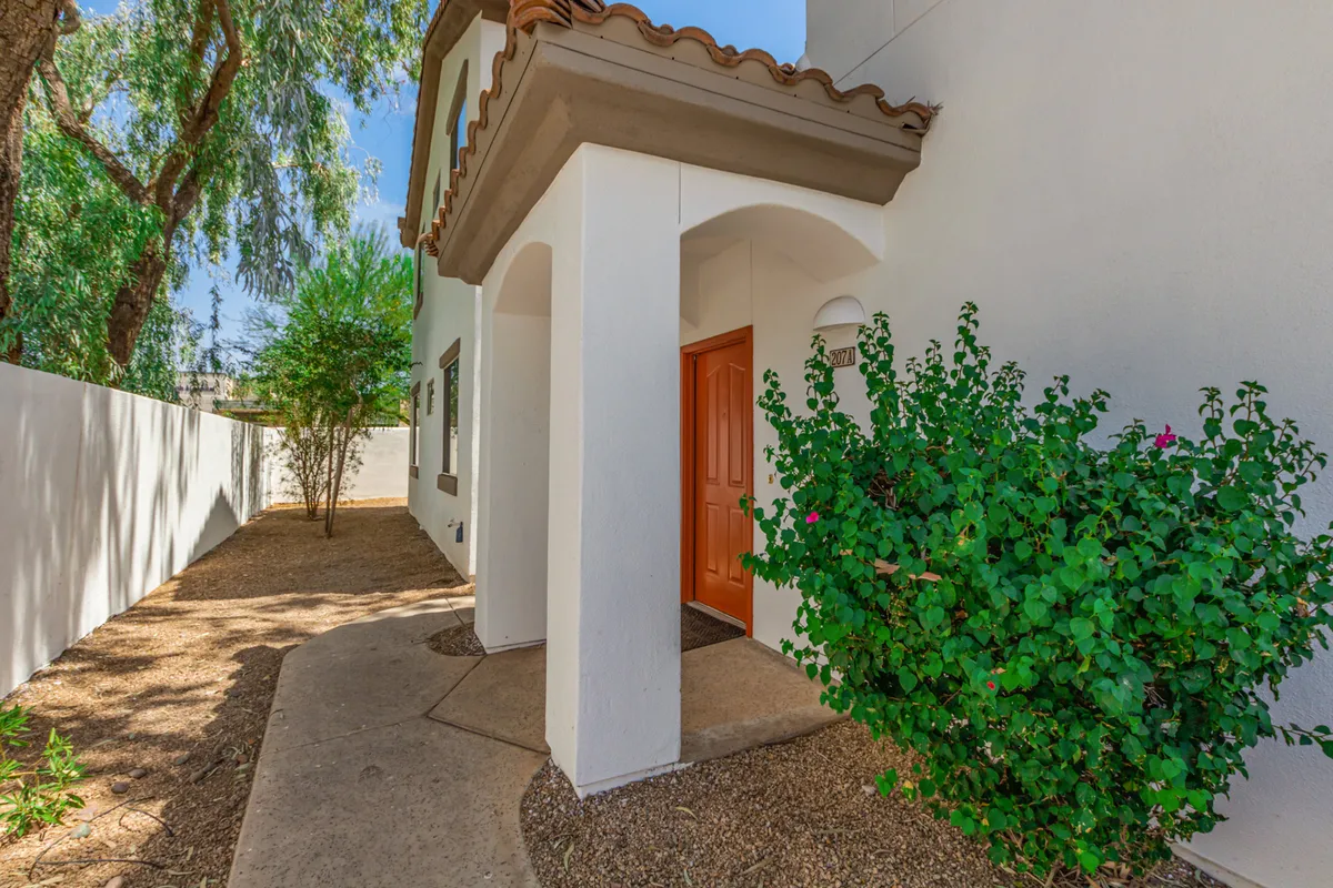 This exterior image showcases a welcoming orange door framed by lush green shrubbery and crisp white walls, suggesting an inviting entryway to the community. The vibrant greenery and outdoor setting evoke the relaxed, sun-drenched lifestyle associated with Arizona living.